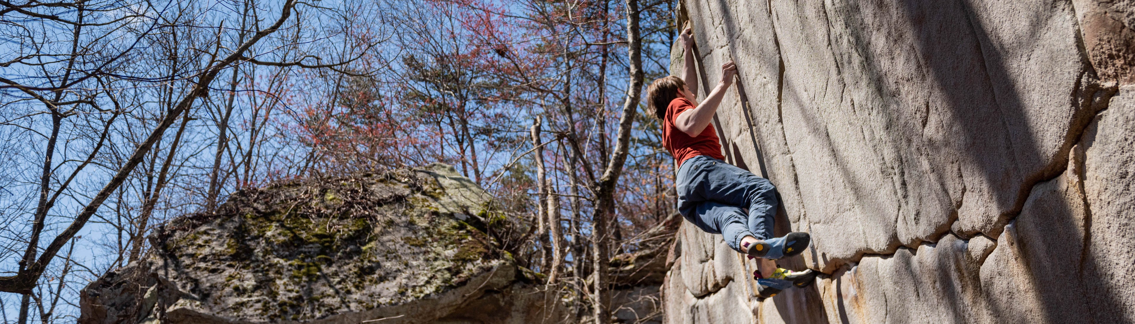 A person wearing a red shirt and climbing shoes scales a large rock wall outdoors on a sunny day, surrounded by leafless trees and boulders.