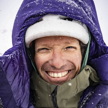 Person wearing a purple winter coat with snow on their face and hood, smiling.