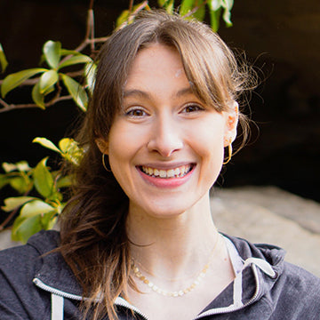 A young woman with long brown hair in a ponytail smiles at the camera. She is wearing a dark zip-up hoodie, gold hoop earrings, and layered necklaces, with green leaves in the background.