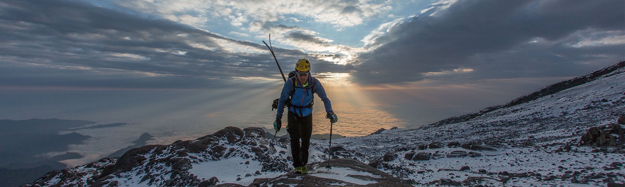 Person hiking on a mountain trail with a scenic sunset or sunrise in the background.