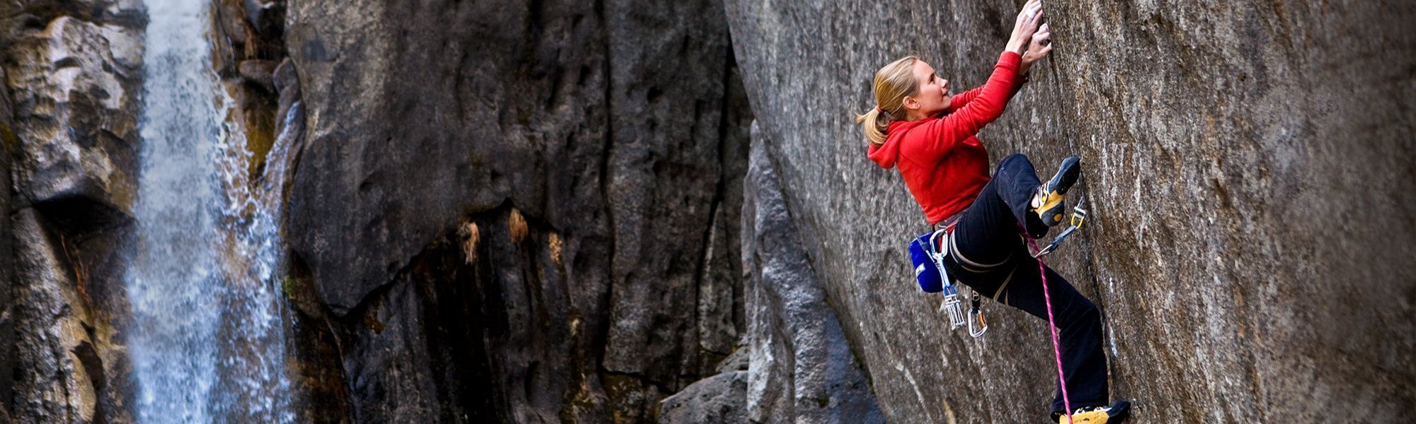 A person wearing a red hoodie and climbing gear is rock climbing on a steep, textured cliff face, with a waterfall and rocky landscape visible in the background.
