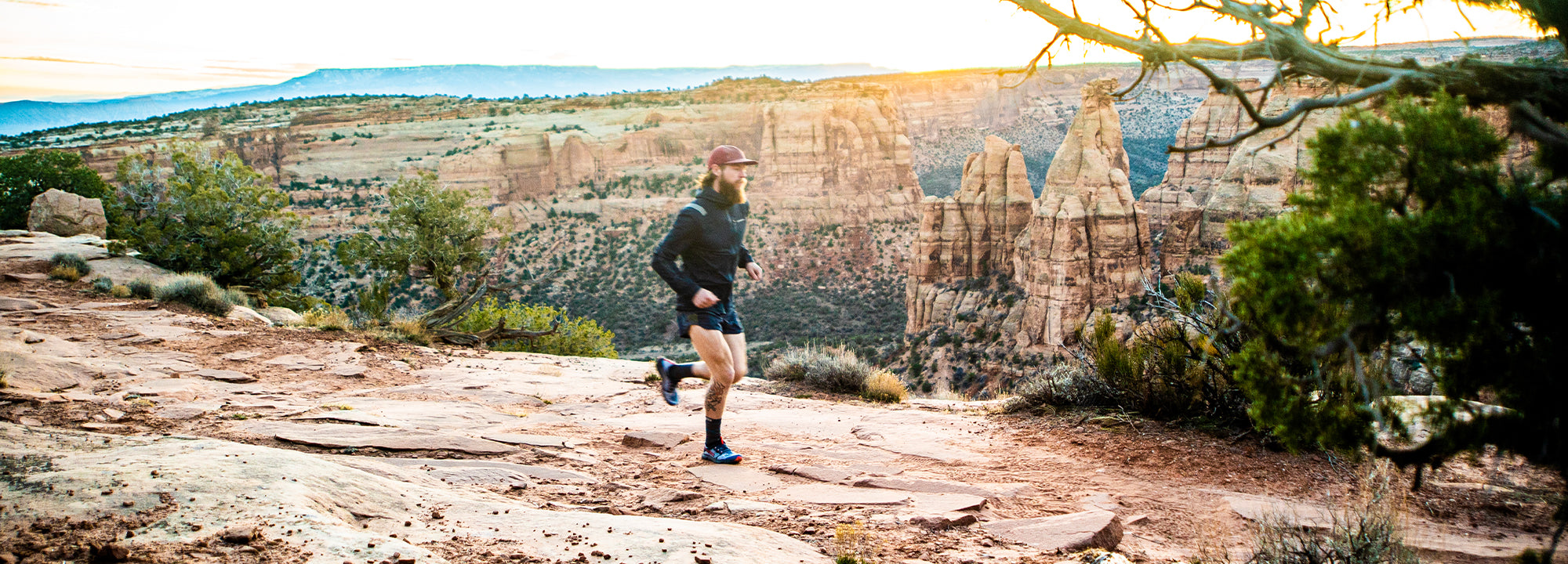 A man wearing a cap, jacket, and shorts runs along a rocky trail on a canyon rim at sunrise, with tall sandstone formations and desert landscape visible in the background.
