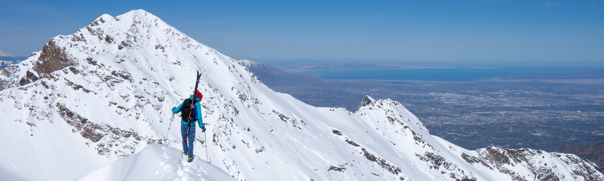 Person standing on a snowy mountain peak with skis, overlooking a vast landscape.