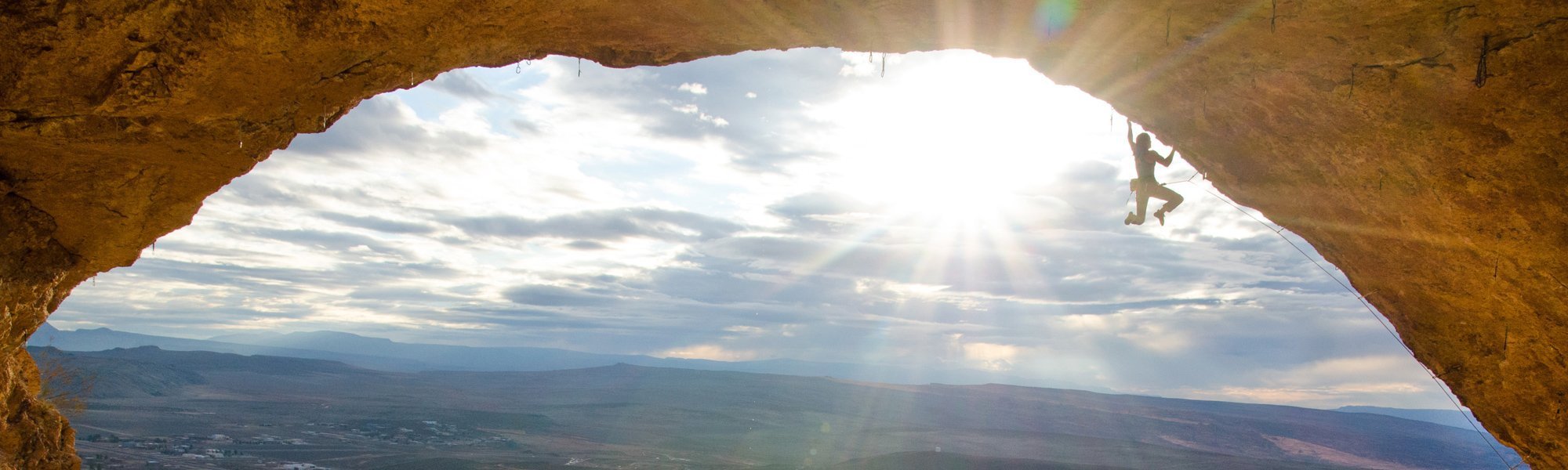A rock climber ascends a cave ceiling, silhouetted against the bright sun and a landscape of distant mountains and a valley under a partly cloudy sky.