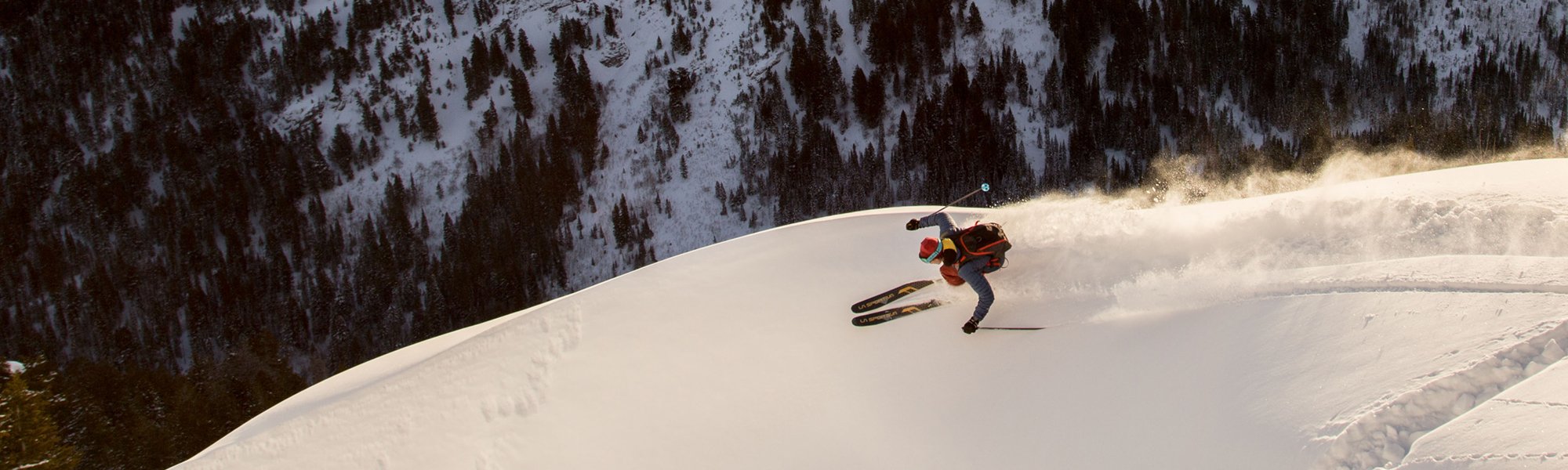 A skier loses balance and falls on a snowy slope, sending up a spray of powder, with a forested mountain hillside in the background.