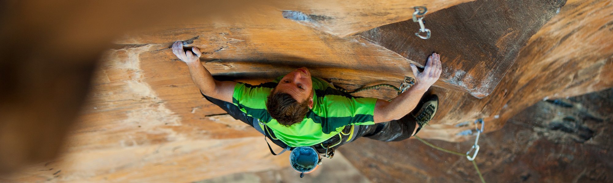 A rock climber wearing a bright green shirt scales a steep, brown rock face, gripping with chalk-covered hands while attached to safety ropes and gear.