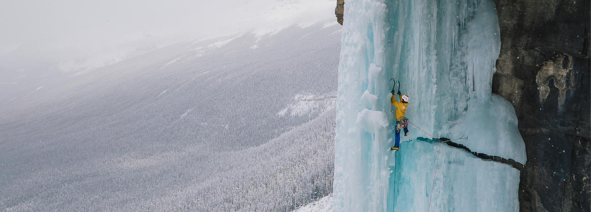 A person in a yellow jacket ice climbs a tall, vertical frozen waterfall on a mountainside, surrounded by snowy, forested landscape and misty weather.
