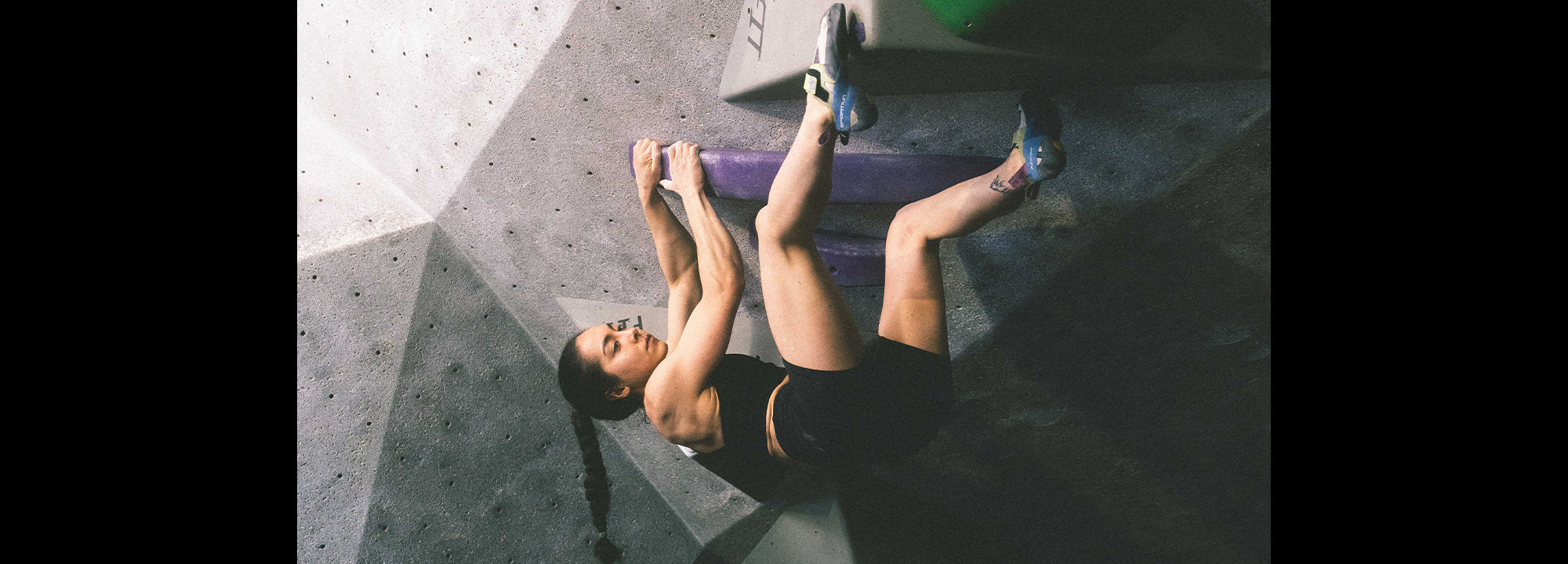 A woman wearing athletic clothes and climbing shoes is bouldering on an indoor climbing wall, gripping purple holds with her hands and feet while leaning back.