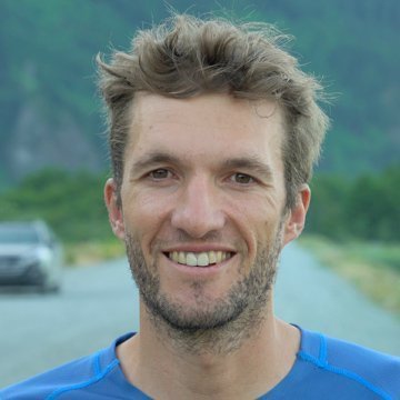Man with a beard and blue shirt standing on a road with greenery in the background