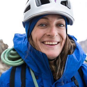 Person wearing a blue jacket and white helmet with a climbing rope, standing against a mountainous background.