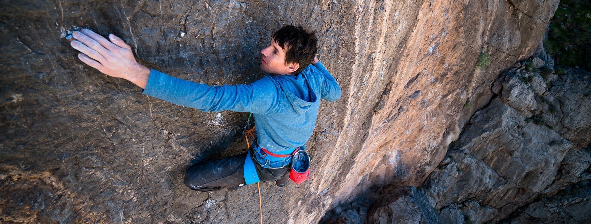 Person climbing a rocky cliff face wearing a blue shirt and red shoes.