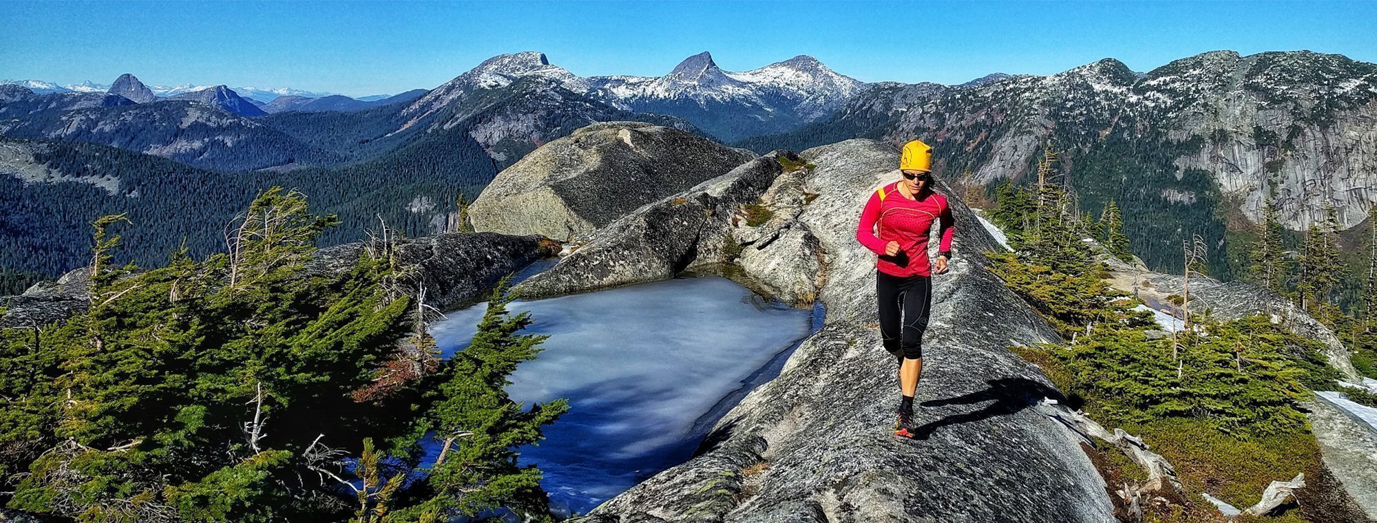A person in a red jacket and yellow hat runs along a rocky mountain ridge beside a small frozen pond, with snow-capped peaks and evergreen trees in the background under a clear blue sky.