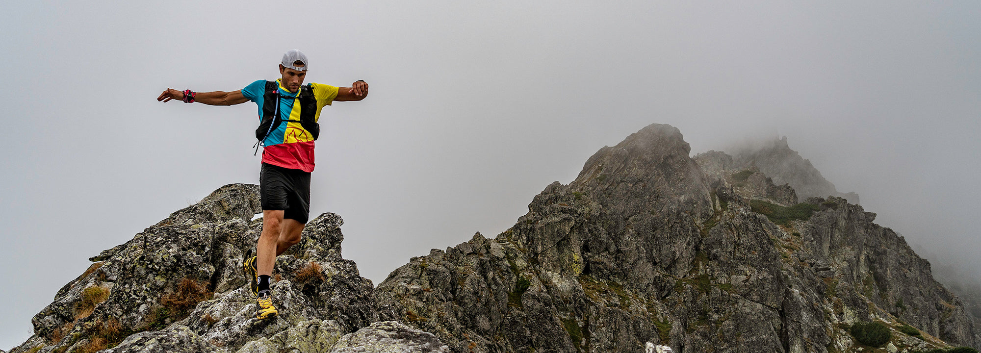 A person in athletic gear and a backpack carefully balances while traversing a rocky mountain ridge under a cloudy, foggy sky. Jagged peaks rise in the background.