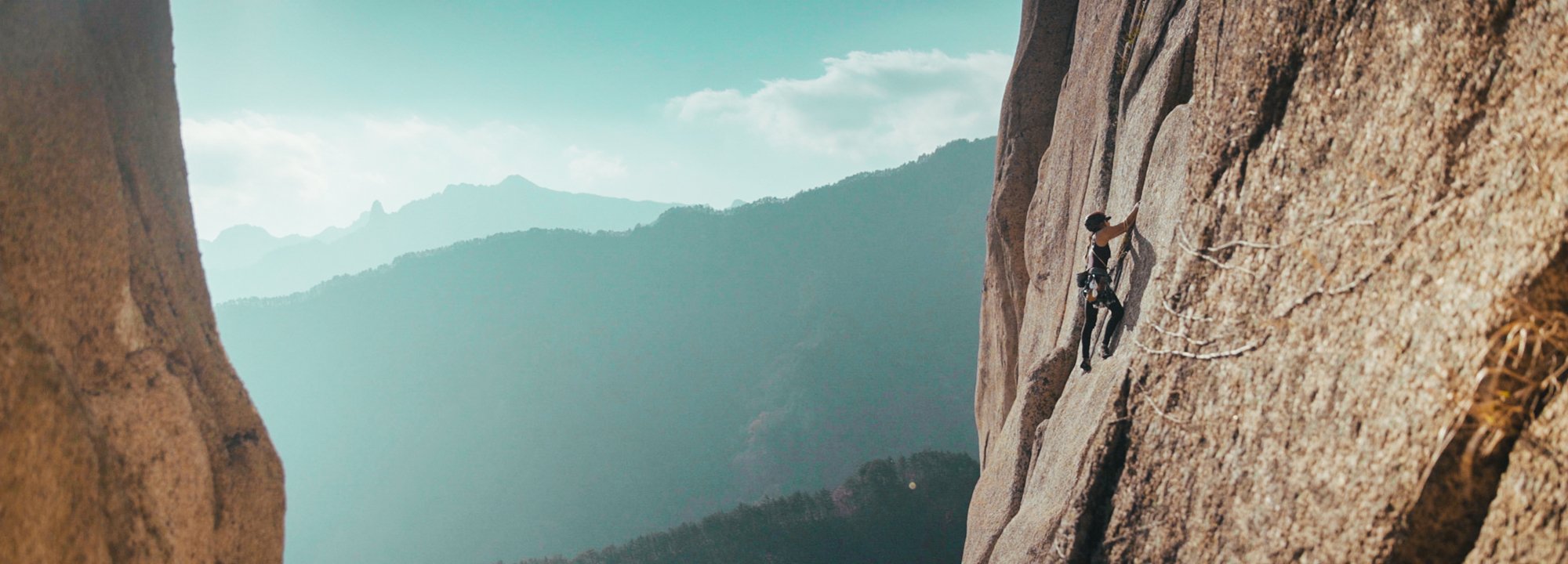 Person climbing a rocky mountain with a scenic view of mountains and sky.