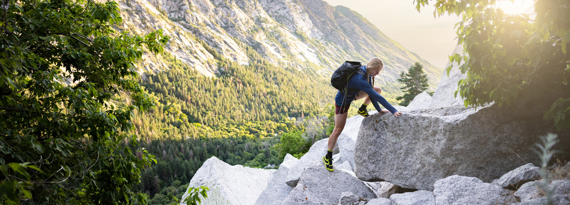 Person hiking on a rocky mountain trail with backpack, surrounded by greenery and mountains.