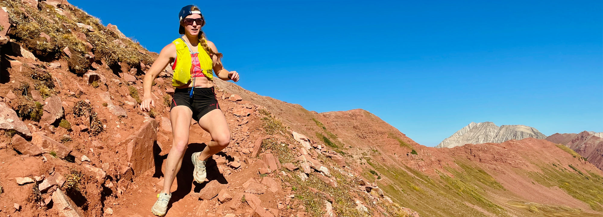 A person wearing a yellow vest and black shorts runs on a rocky mountain trail under a clear blue sky, with red and green hills and distant peaks in the background.