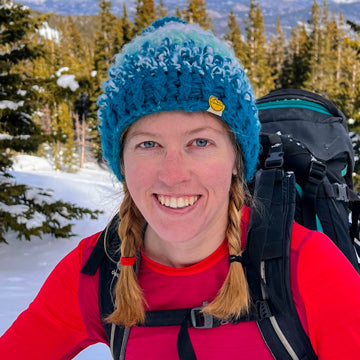 Person wearing a blue knit hat and red jacket with a backpack in a snowy landscape.