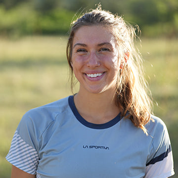 A woman with long brown hair in a ponytail smiles outdoors. She wears a light blue athletic shirt with La Sportiva printed on it. The background is grassy and sunlit.