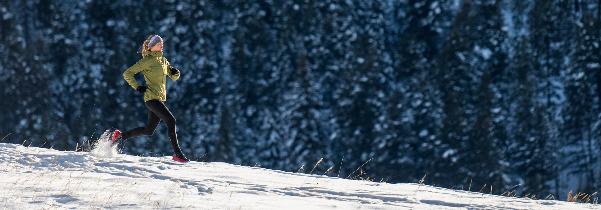 A person in a green jacket and black leggings runs through snow on a sunlit hillside, with a forest of dark, snow-covered trees in the background.