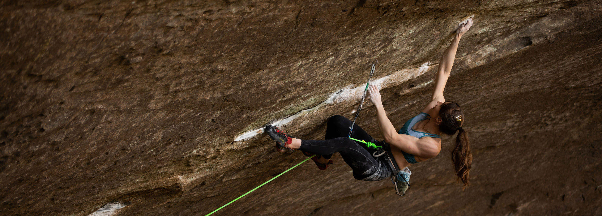 A woman in athletic clothes climbs a steep rock face, reaching with her right hand for a hold while secured by a green climbing rope and quickdraws.