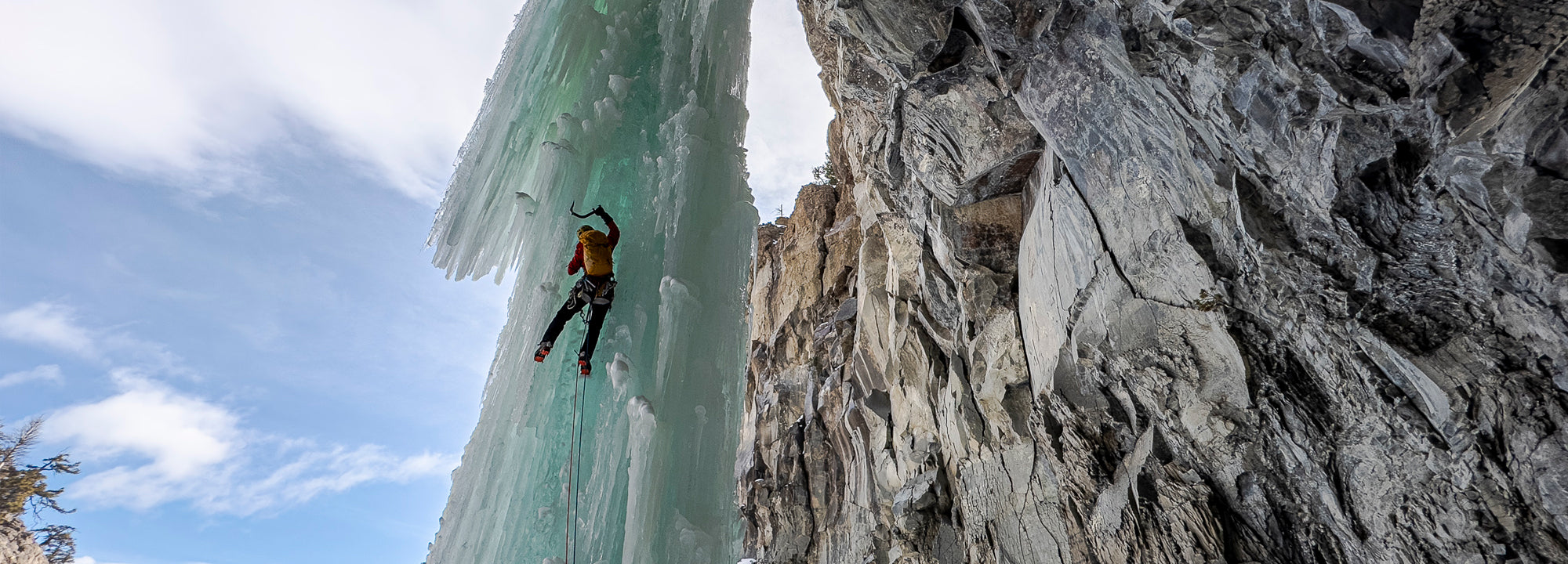 A person wearing a helmet and winter gear climbs a tall, vertical icicle formation beside a rugged rock cliff under a partly cloudy sky.