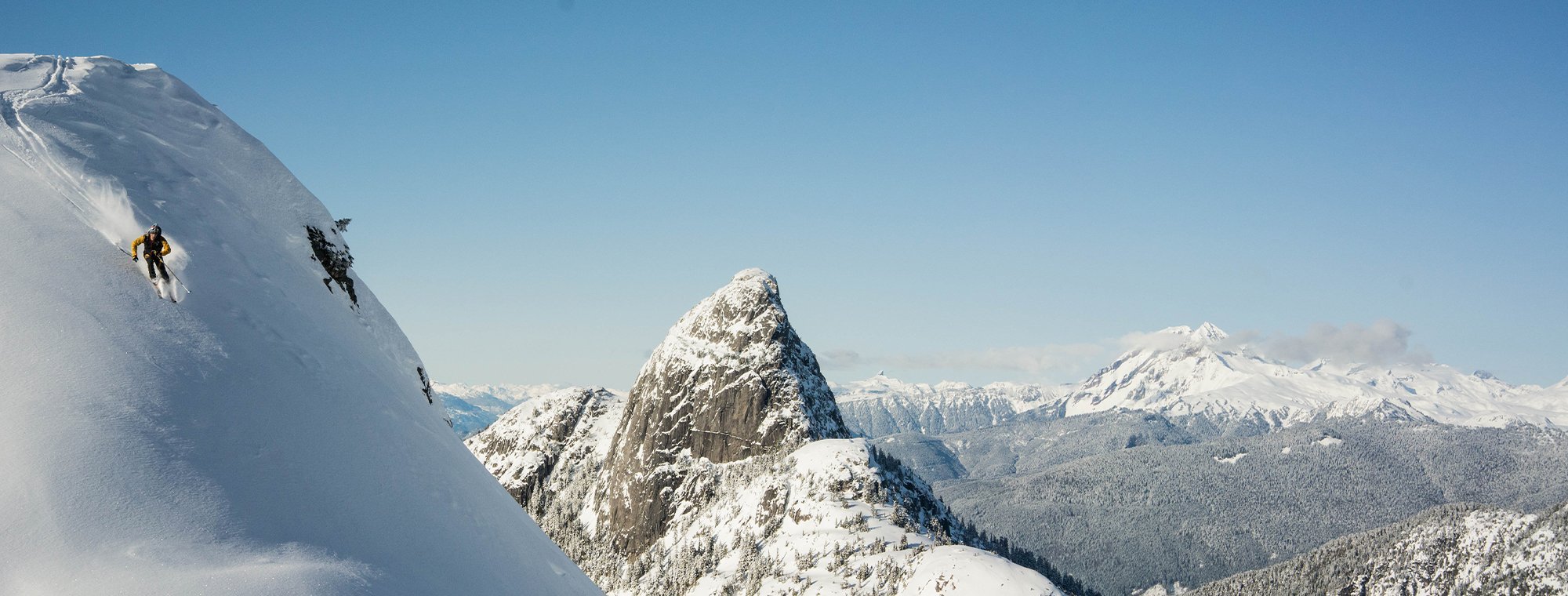 A skier descends a steep, snow-covered mountain slope with a backdrop of jagged, snow-capped peaks and a clear blue sky.