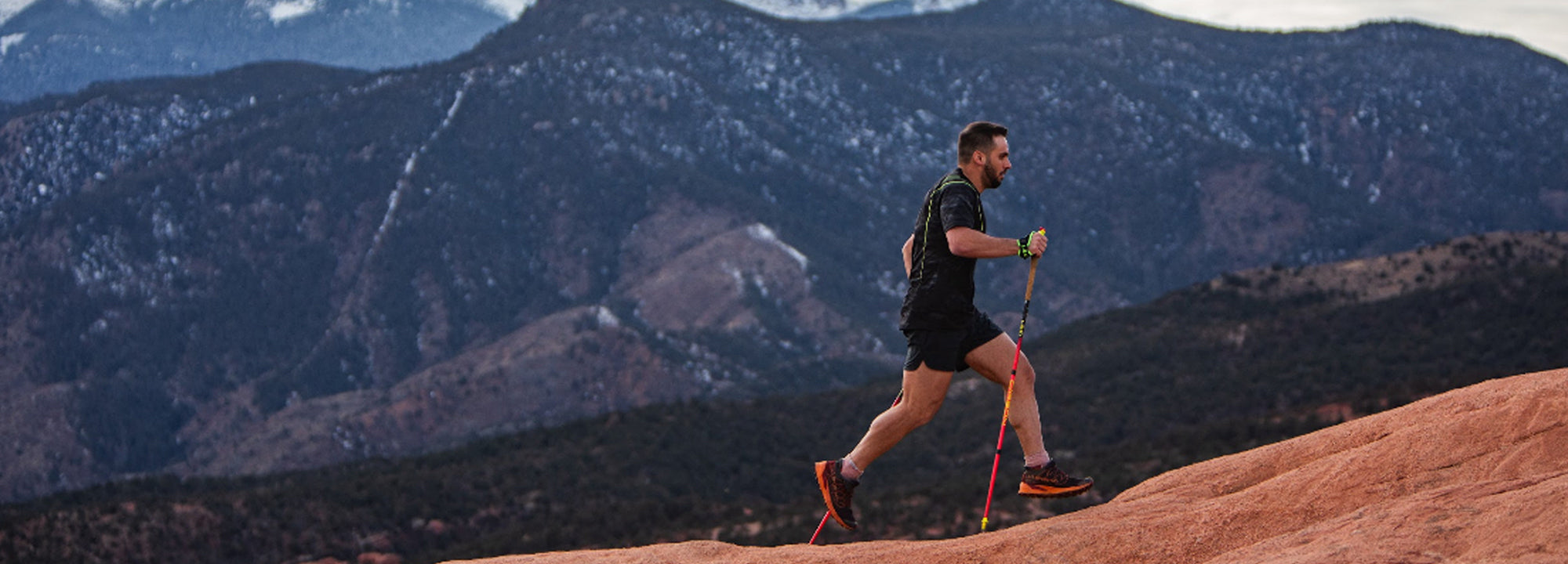 A man in athletic gear hikes uphill on a rocky trail using trekking poles, with forested mountains and snow-dusted peaks in the background.