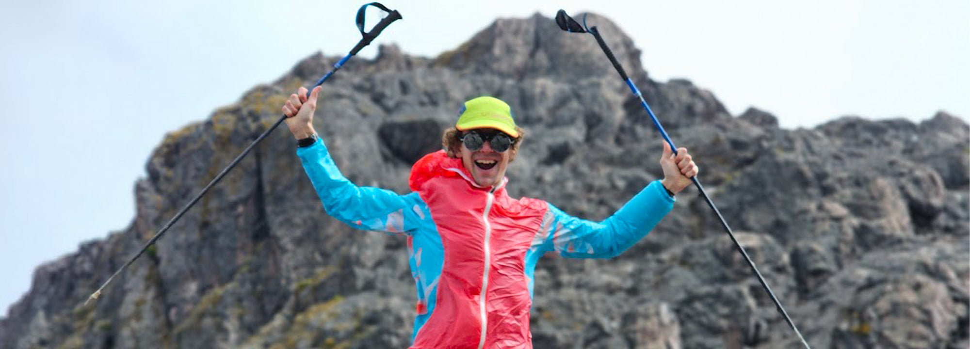 A smiling hiker wearing a bright jacket, neon green cap, and sunglasses raises trekking poles triumphantly with a rocky mountain in the background.