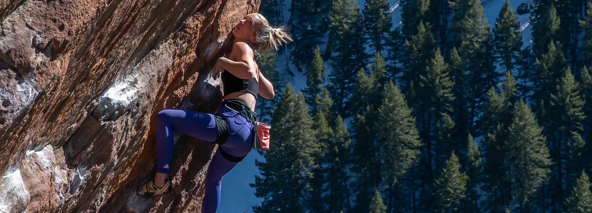 A woman in athletic clothing climbs a steep, rocky cliff with a forest of tall evergreen trees and snow patches visible in the background. She grips the rock with determination under clear daylight.