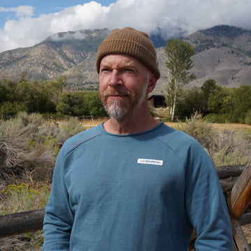 Man wearing a blue shirt and brown beanie standing in a mountainous landscape.