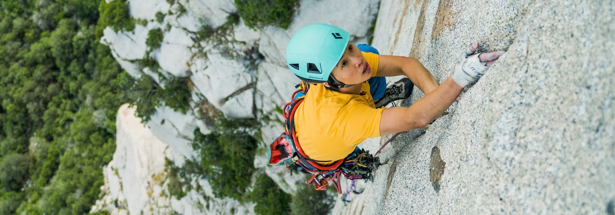 A climber wearing a turquoise helmet and yellow shirt ascends a steep rock face, gripping the stone with chalked hands. Forested cliffs are visible in the background.