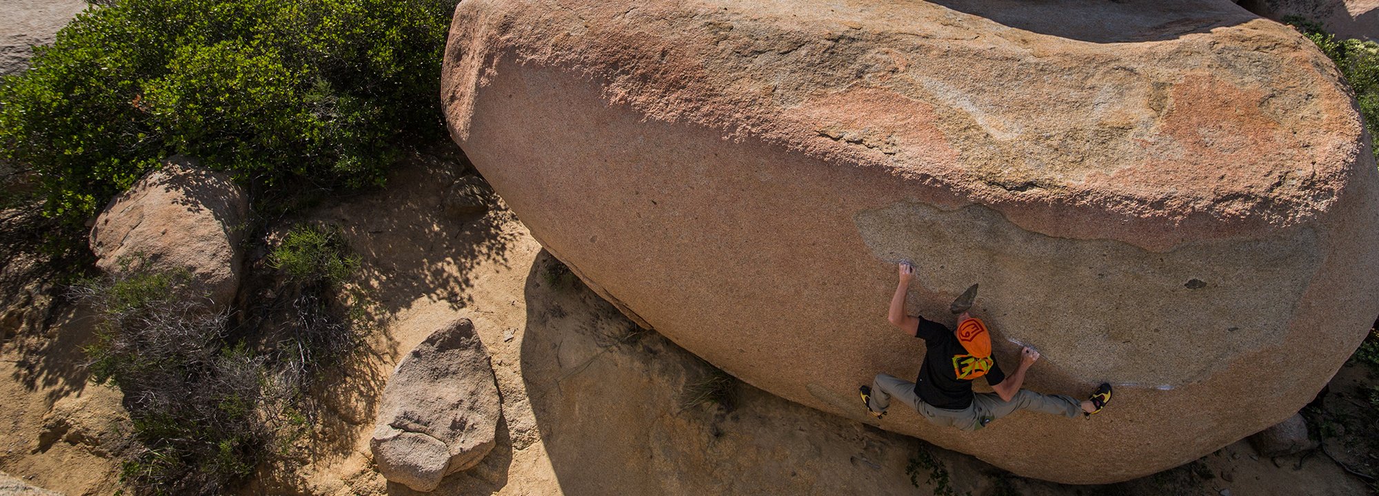 A climber wearing a black shirt and orange helmet scales a large, rounded boulder in an outdoor, rocky landscape with sparse vegetation.