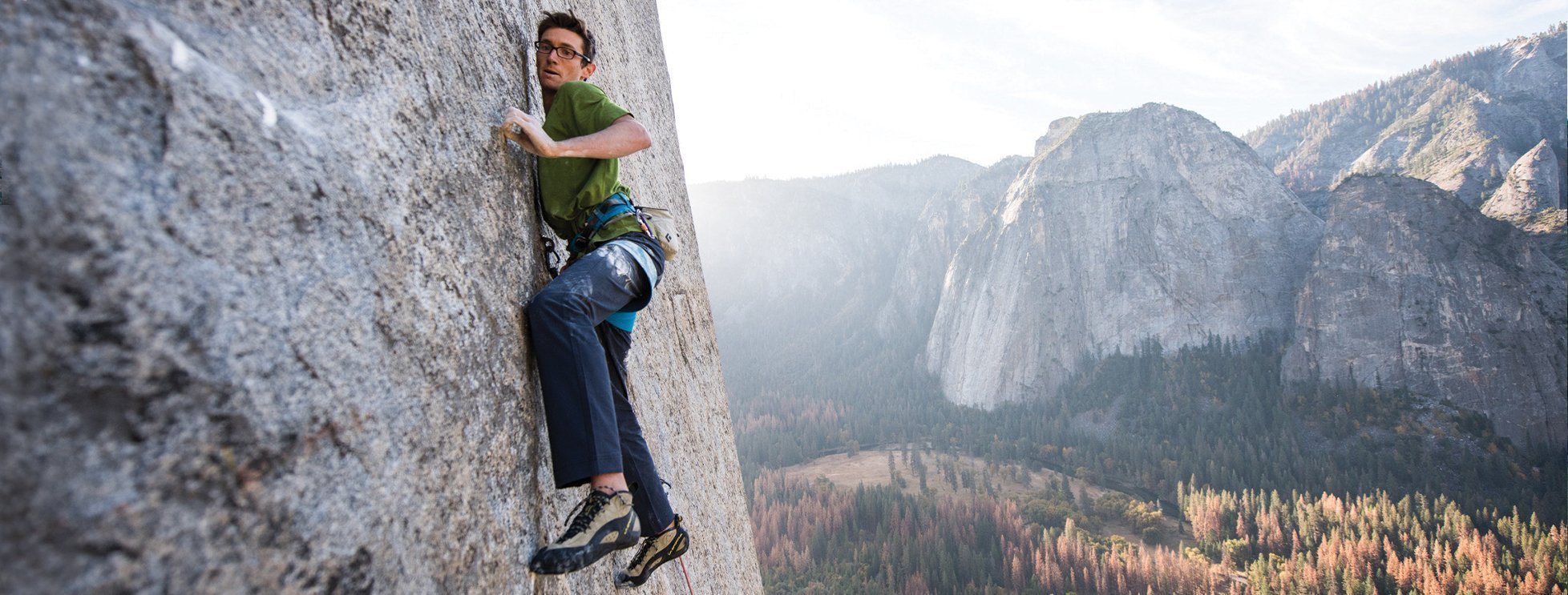 A rock climber wearing a green shirt and blue pants ascends a steep cliff face with a harness, while a scenic mountain and forest landscape stretches out in the background.