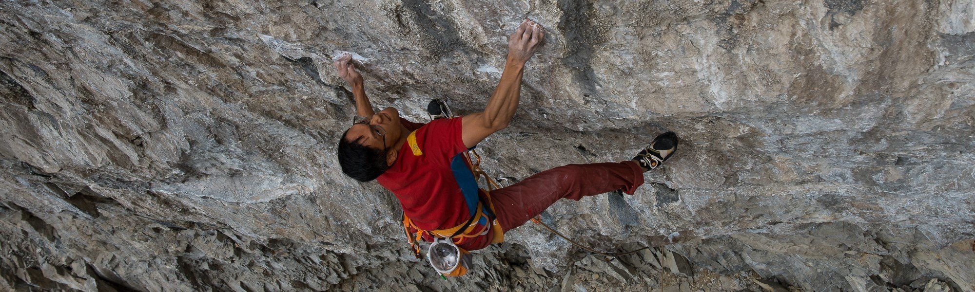 A rock climber wearing a red shirt, harness, and climbing shoes ascends a steep, rocky overhang, gripping small holds with both hands and feet against a rough, textured cliff face.