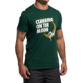 A man stands relaxed in a green La Sportiva Moon Rock T-Shirt Men's (2025), featuring "CLIMBING ON THE MOON" and a climber scaling a crescent moon.