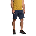 Un homme vêtu d'un T-shirt jaune, d'un short La Sportiva Roots Men's (2025) bleu marine et de chaussures de randonnée noires avec des accents orange se tient debout sur un fond blanc uni. Son visage n'est pas visible.