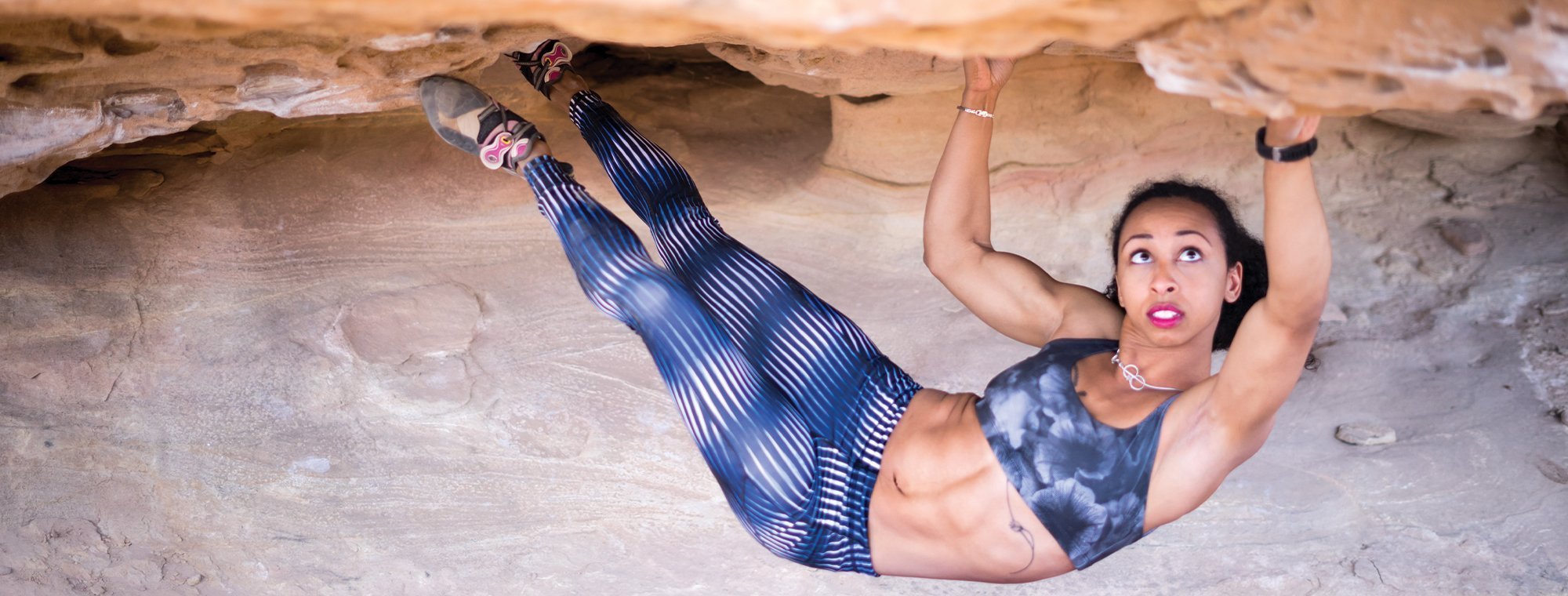 A woman in athletic clothing climbs horizontally on a rock ceiling, gripping the rock with her hands and feet. She appears focused and strong, with toned muscles visible. The environment is rocky and outdoors.