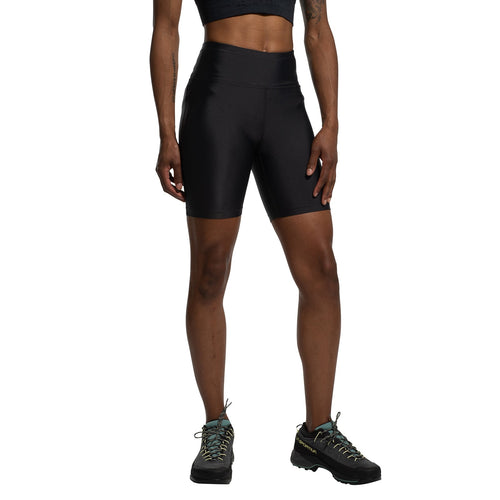 Une personne se tient debout sur un fond blanc, les bras détendus, vêtue d'un soutien-gorge de sport noir, d'un short La Sportiva Aerial Shorts Women's (2025) noir à soufflet d'entrejambe et taille haute, et de chaussures de randonnée noires.