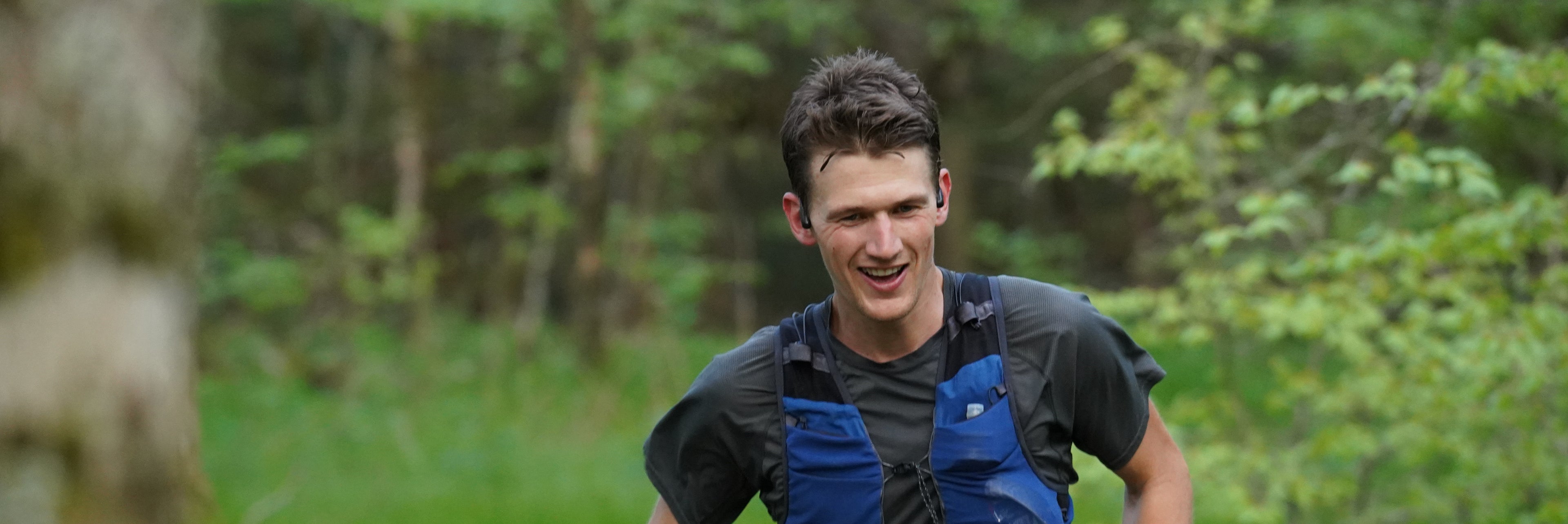 A young man in a blue vest and black shirt runs through a green forest, smiling and appearing energetic.