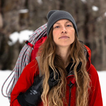 Person in a red jacket and gray beanie with climbing gear, standing in a snowy forest.