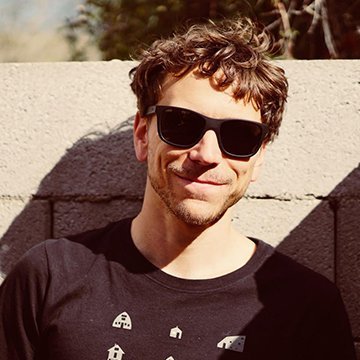 Man wearing sunglasses and a black t-shirt with a logo, standing against a stone wall.