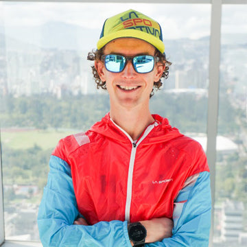 Person wearing a red jacket and yellow cap with sunglasses, standing in front of a window with a scenic view.