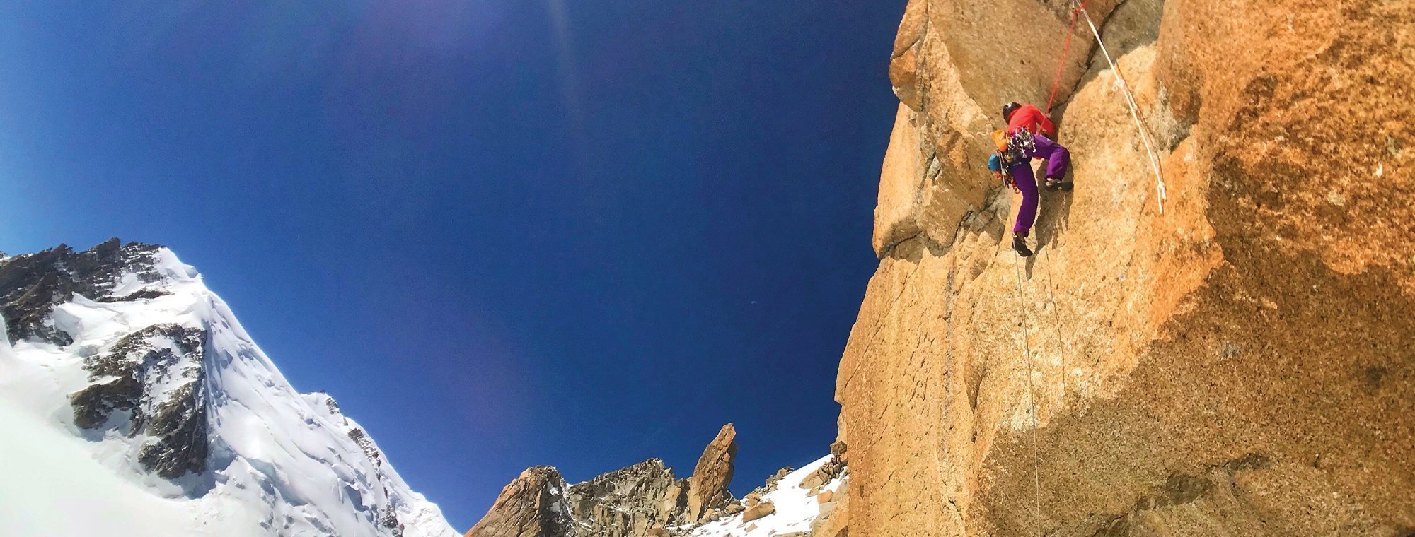 Person climbing a rocky mountain face with snow-capped peaks in the background