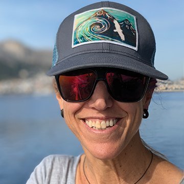 Person wearing a cap with a wave design and sunglasses, standing by a body of water.