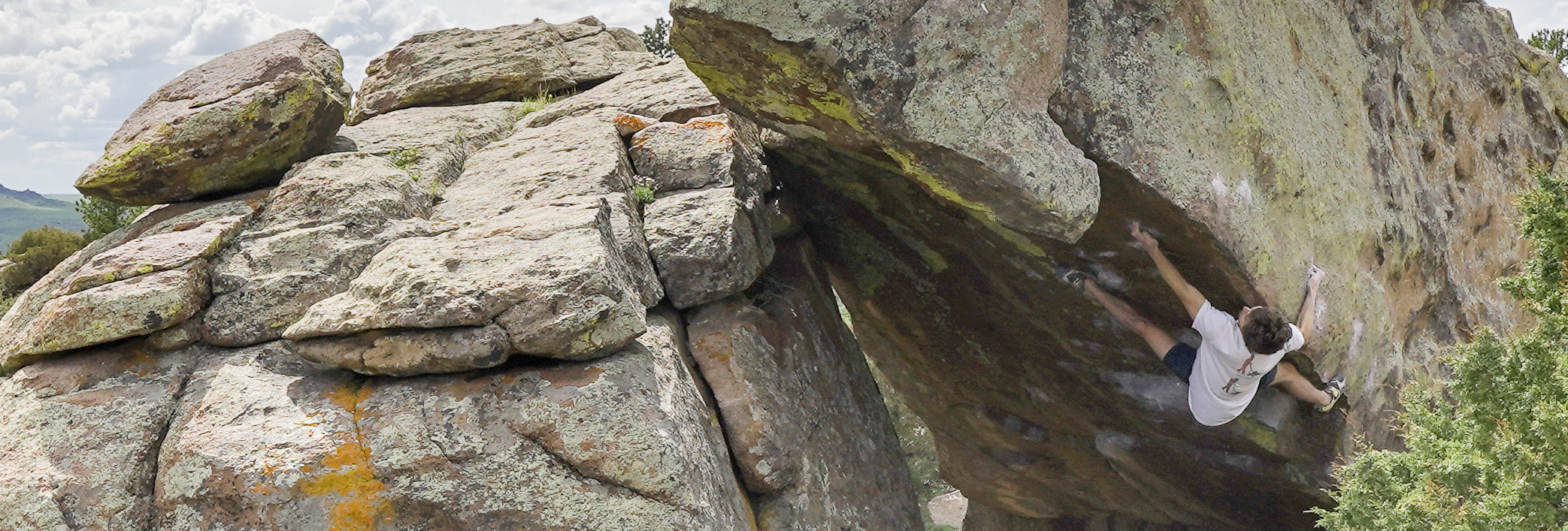 A person rock climbing on a large, overhanging boulder with rough, textured surfaces, surrounded by greenery and a partly cloudy sky.