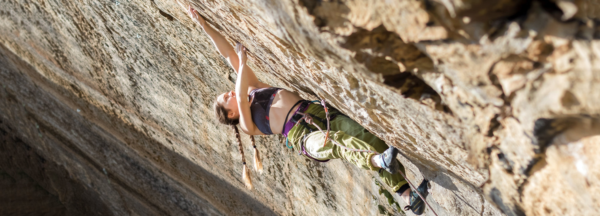 A rock climber with long hair, wearing a sports bra and green pants, climbs a steep, textured rock face outdoors, reaching upward with one arm while secured by climbing gear.