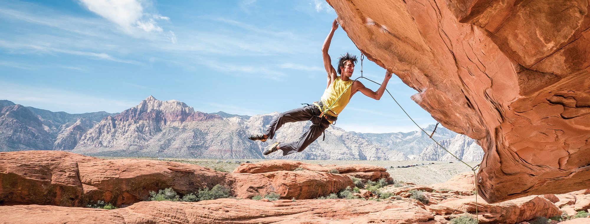 A climber in a yellow shirt hangs from a steep red rock overhang, with a mountainous desert landscape and blue sky in the background.