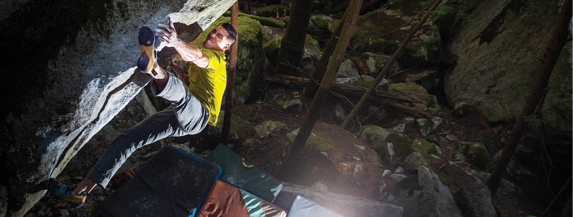 A climber in a yellow shirt and gray pants grips a rocky overhang in a forest, with crash pads below for safety and sunlight filtering through the trees.