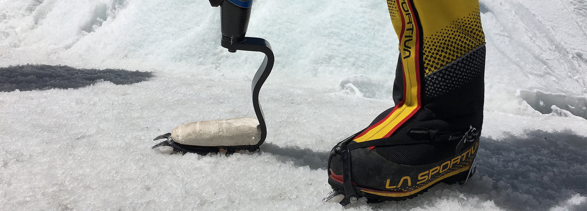 A person with a prosthetic leg and a yellow La Sportiva mountaineering boot stands on snow; the prosthetic foot is equipped with a crampon for traction.