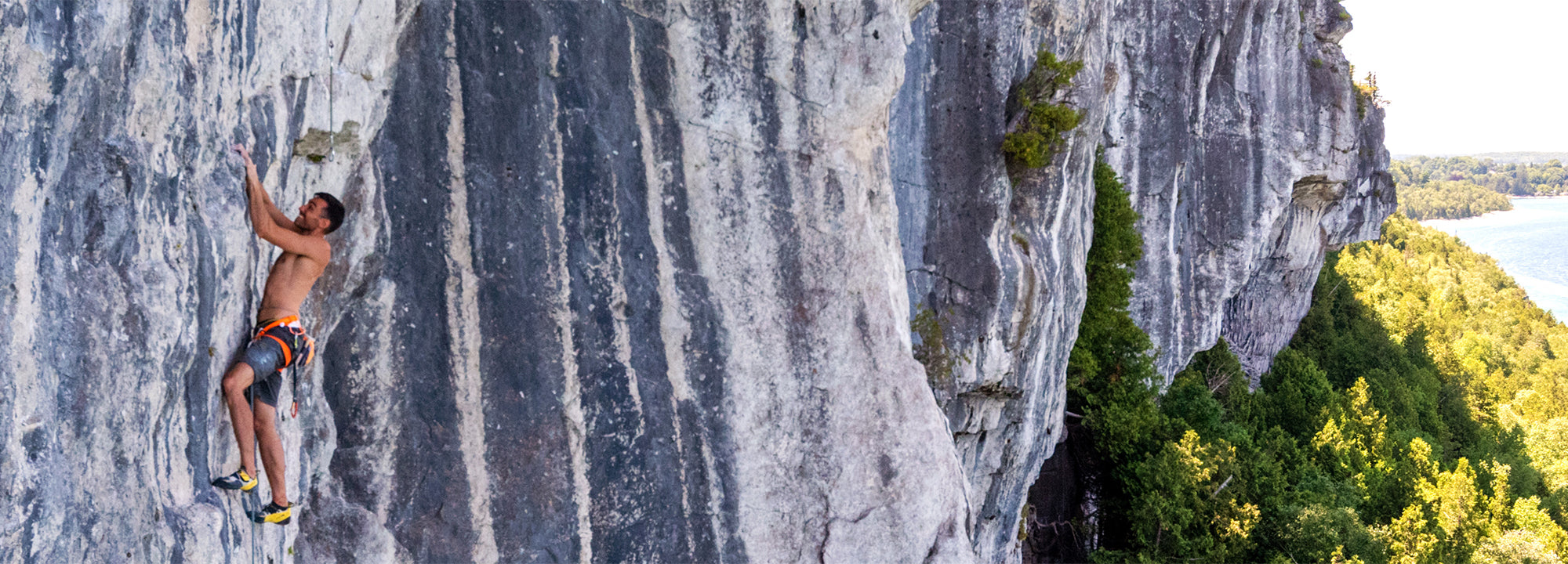 A person wearing a harness climbs a steep, rocky cliff face above a forested area and water, grasping for holds on the rock wall.
